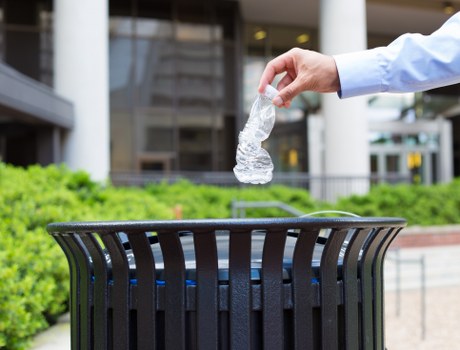 A person's hand, dressed in a light blue shirt, is seen dropping a crumpled clear plastic water bottle into a black metal rubbish bin with vertical slats, situated outdoors in an urban environment. In the background, there are modern office buildings with large glass windows, metal railings, and a paved walkway. The scene is well-lit with natural daylight, and the waste bin is positioned on a paved surface next to a curb or pathway, consistent with a commercial or business area in Acton. The hand appears to be releasing the plastic bottle over the open top of the bin, emphasizing waste disposal activities typical of rubbish removal services provided by Office Clearance Acton. The environment indicates a clean, organized city space where rubbish collection and recycling efforts are ongoing.