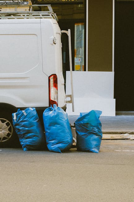A large, bright blue plastic wheelie bin with a closed lid is positioned on the pavement beside a white delivery van with a metal door featuring a vertical black handle. The bin shows signs of dirt an