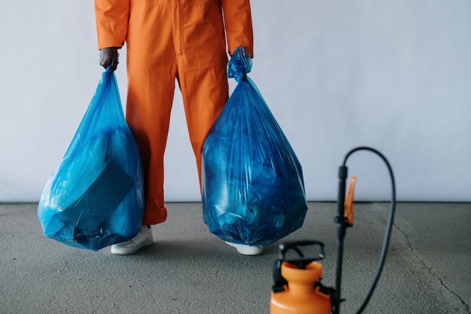 A person wearing orange overalls and white shoes is standing indoors on a grey concrete floor, holding two large blue rubbish bags filled with waste, with one bag in each hand. The bags appear to be m